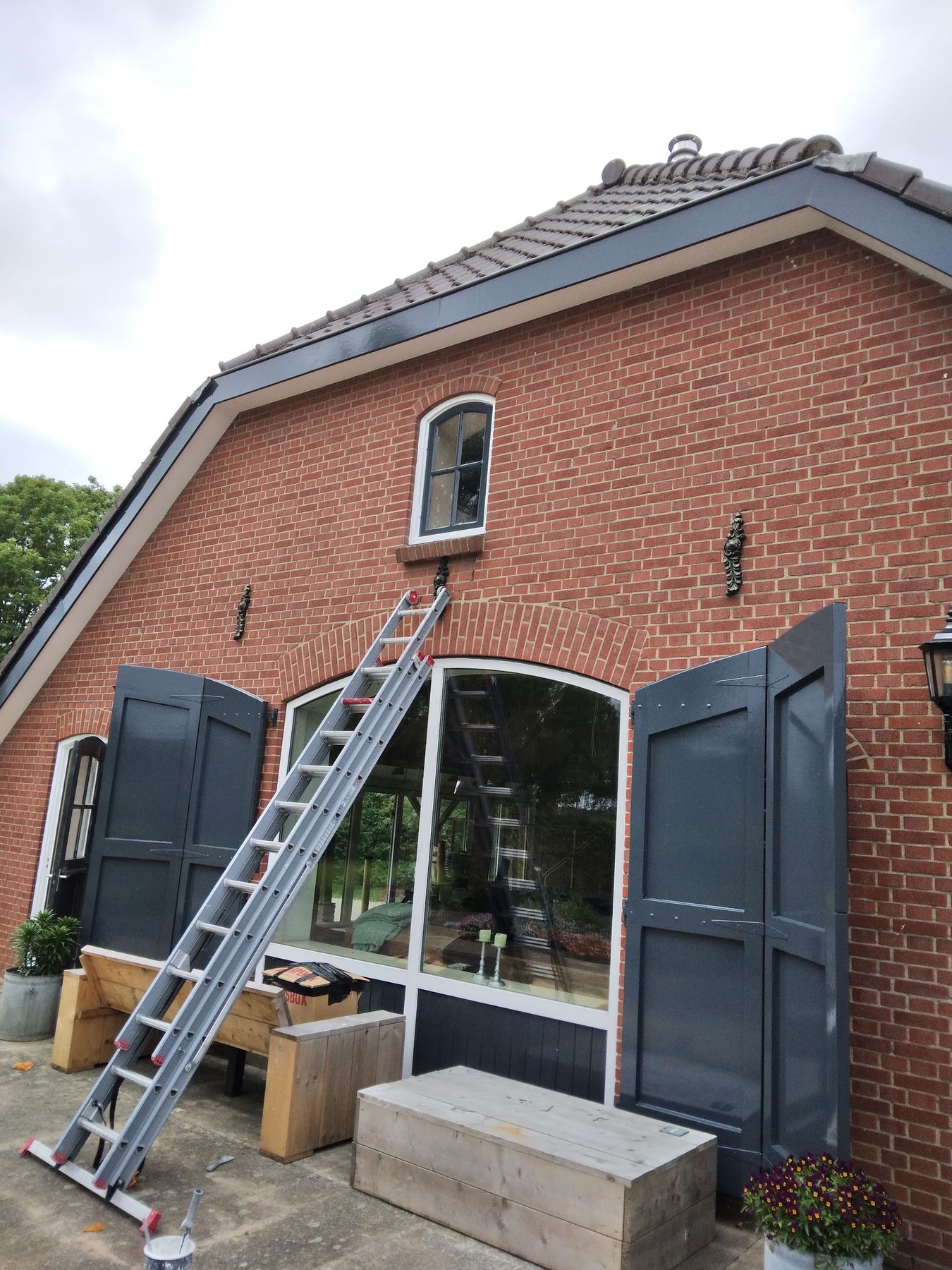 Topgevel van een bakstenen woning met donkergrijze luiken en kozijnen, ladder tegen de gevel